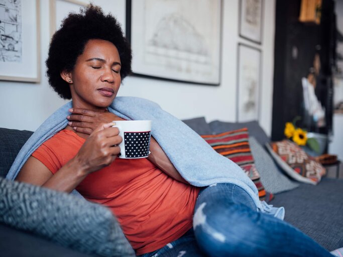 Reife Frau sitzt krank mit Halsschmerzen auf der Couch und trinkt Tee | © gettyimages.de | Brothers91