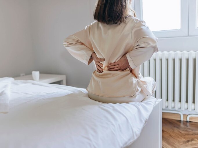 Frau von hinten auf dem Bett sitzend, hat Schmerzen im Rücken | © gettyimages.de | PixelsEffect