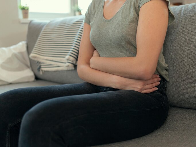 Frau sitzt mit Magen-Darm-Schmerzen auf der Couch und hält sich den Bauch | © gettyimages.de | PixelsEffect