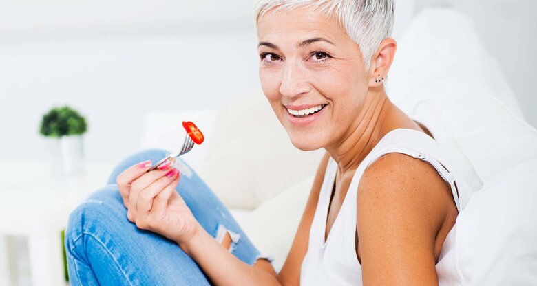 Hübsche, ältere Frau isst einen Salat auf dem Sofa | © iStock | Vesnaandjic