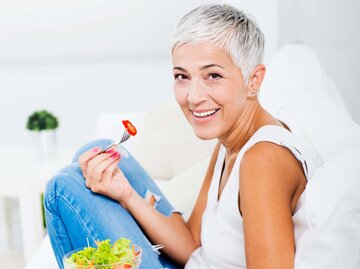 Hübsche, ältere Frau isst einen Salat auf dem Sofa | © iStock | Vesnaandjic