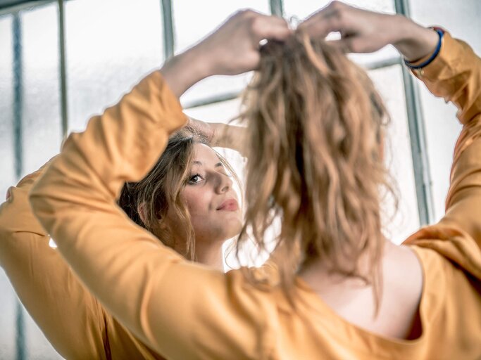 Frau mit hellbraunem, gewelltem Haar bindet sich vor dem Spiegel einen Pferdeschwanz | © iStock | Ababsolutum