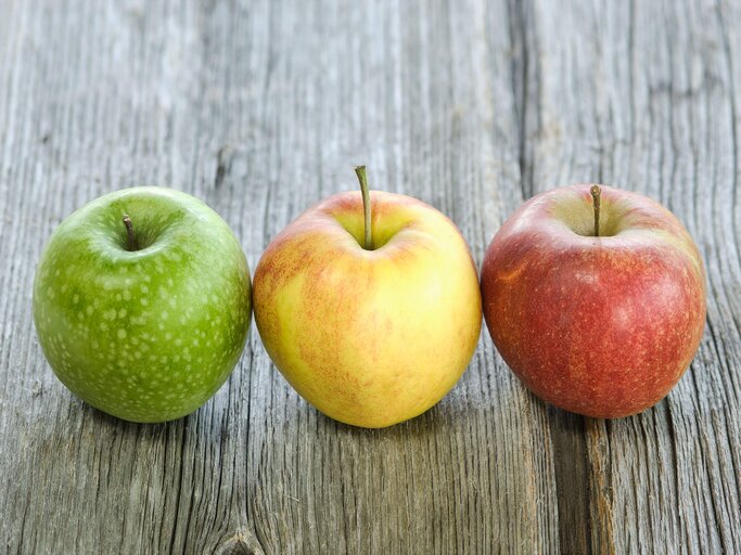 Grüner, gelber und roter Apfel liegen nebeneinander auf einer Holzablage | © iStock | Tuned_In