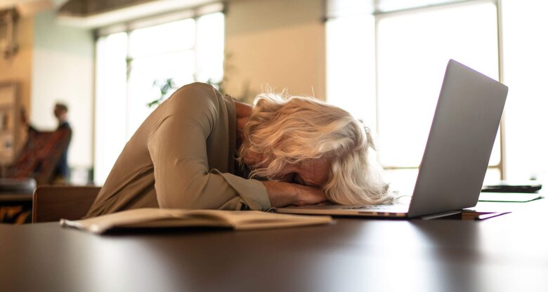Reife Frau mit grauen halblangen Haaren schläft am Schreibtisch vor ihrem Computer. | © iStock | FG Trade