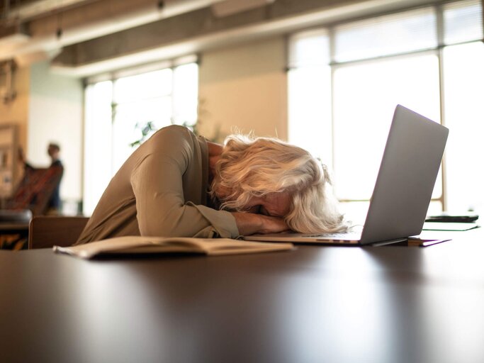 Reife Frau mit grauen halblangen Haaren schläft am Schreibtisch vor ihrem Computer. | © iStock | FG Trade