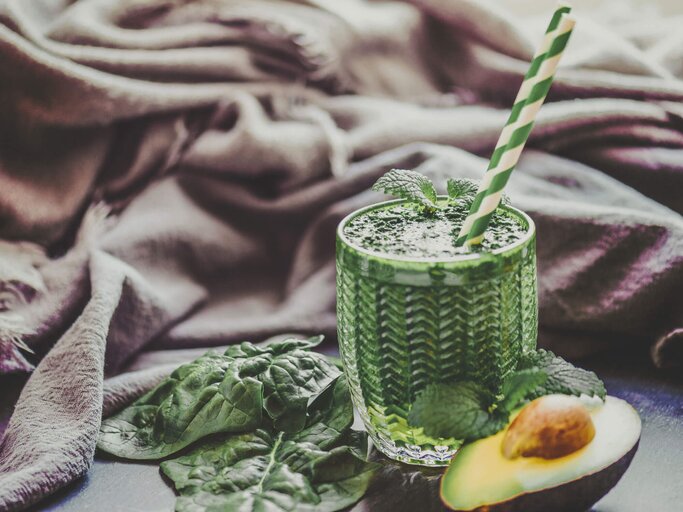 Glas mit grünem Smoothie und Strohhalm, daneben eine aufgeschnittene Avocado. | © iStock | Rocky89