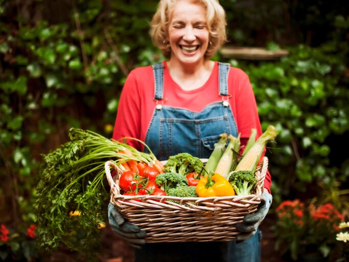 Ältere Frau steht im Garten und hält glücklich einen Korb mit Gemüse. | © iStock.com / stevecoleimages