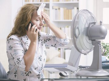 Frau sitzt am Schreibtisch vor einem Ventilator und leidet unter Hitzewallungen. | © iStock.com / humonia