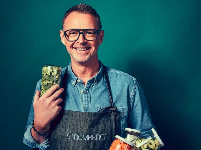 Sternekoch Holger Stromberg vor einem blauen Hintergrund und Gläsern mit Lebensmitteln in der Hand.