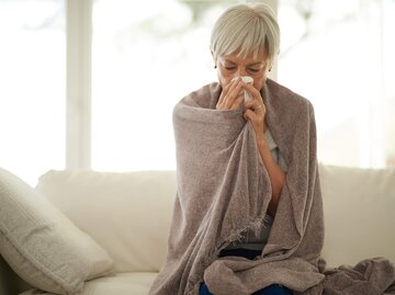 Reife Frau sitzt mit einer Decke auf der Couch und hat einen chronischen Schnupfen / Dauerschnupfen. | © iStock.com / Cecilie_Arcurs