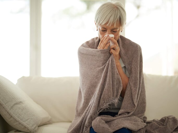 Reife Frau sitzt mit einer Decke auf der Couch und hat einen chronischen Schnupfen / Dauerschnupfen. | © iStock.com / Cecilie_Arcurs