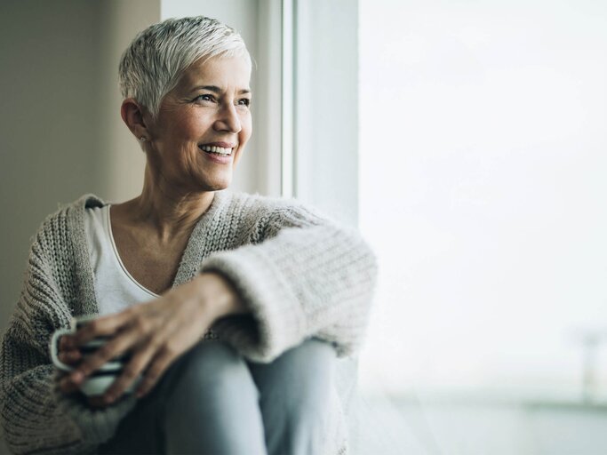 Frau mit grauen kuzen Haaren sitzt am Fensterbrett und schaut in die Ferne. | © iStock.com / skynesher