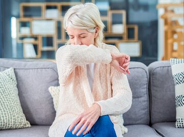 Reife Frau sitzt auf dem Sofa und hustet sich in die Armbeuge | © gettyimages.de | Moyo Studio