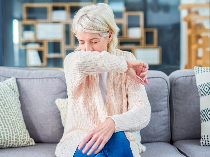 Reife Frau sitzt auf dem Sofa und hustet sich in die Armbeuge | © gettyimages.de | Moyo Studio