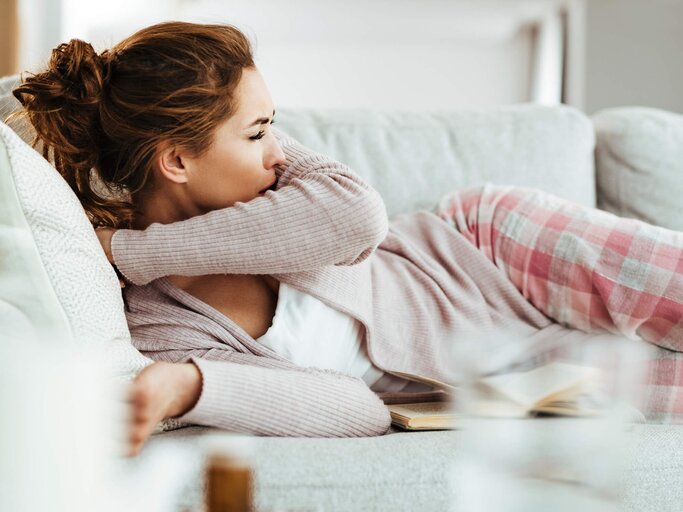 Frau liegt auf dem Sofa und hustet in die Armbeuge | © gettyimages.de | Drazen Zigic