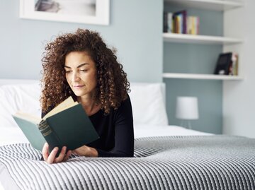 Frau liegt auf ihrem Bett und liest entspannt ein Buch | © Getty Images/	Justin Lambert