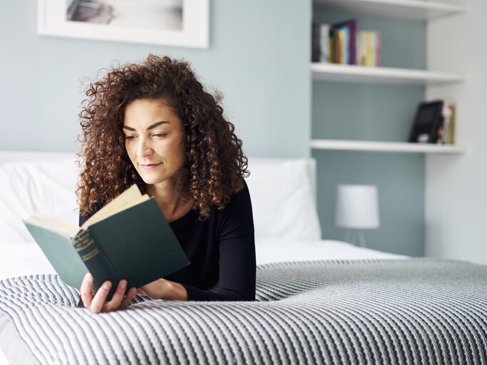 Frau liegt auf ihrem Bett und liest entspannt ein Buch | © Getty Images/	Justin Lambert