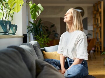 Frau sitzt auf dem Sofa und blickt entspannt in die Sonne | © Getty Images/BartekSzewczyk