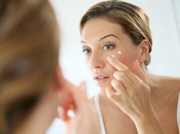 Eine Frau schaut in den Spiegel und verteilt Creme unter ihrem Auge | © Adobe Stock/goodluz