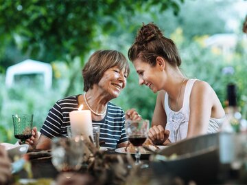 Mutter und Tochter sitzen im Sommer gemeinsam am Essenstisch im Garten und unterhalten sich. | © Getty Images / Hinterhaus Productions