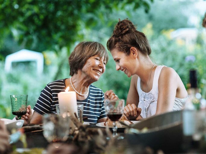 Mutter und Tochter sitzen im Sommer gemeinsam am Essenstisch im Garten und unterhalten sich. | © Getty Images / Hinterhaus Productions