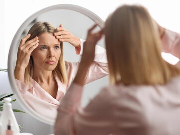 Frau in mittlerem Alter blickt in den Spiegel und betrachet ihre Falten | © Adobe Stock/Prostock-studio