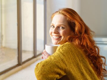 Rothaarige Frau sitzt lächelnd am Fenster | © AdobeStock/contrastwerkstatt